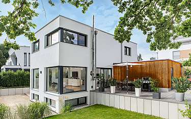 Modernes weißes Haus mit großer Holzterrasse, Garten und Bäumen im Vordergrund.