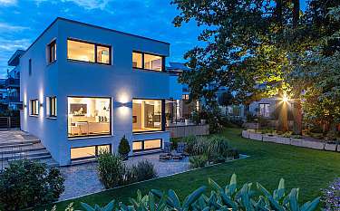Modernes zweistöckiges Haus mit beleuchteten Fenstern und gepflegtem Garten bei Dämmerung.