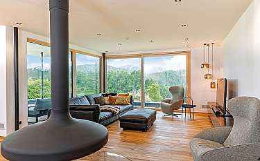 Modernes Wohnzimmer mit Kamin, Sofa, Sessel und großen Fenstern zum grünen Tal.