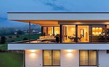 Moderne Villa mit beleuchtetem Innenraum und Panoramafenstern über grüner Landschaft.