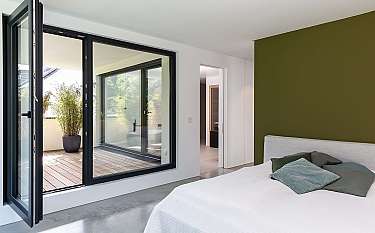 Helles Schlafzimmer mit offener Balkontür, großer Fensterfront und grünem Akzent an der Wand.