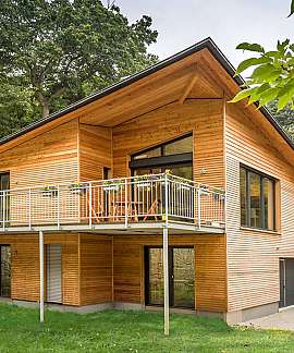 Zweistöckiges Holzhaus mit Balkon und großer Fensterfront in bewaldeter Umgebung.