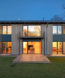 Modernes zweistöckiges Holzhaus mit beleuchteten Fenstern, Terrasse und Balkon im Garten.