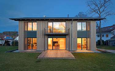Modernes zweigeschossiges Holzhaus mit großen Fenstern und Terrasse im Abendlicht.