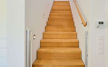 Holztreppe mit Wandbeleuchtung, Handlauf und weißen Wänden in einem modernen Haus.