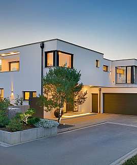 Modernes weißes Einfamilienhaus mit beleuchteter Fassade, Garten und Doppelgarage bei Abenddämmerung.