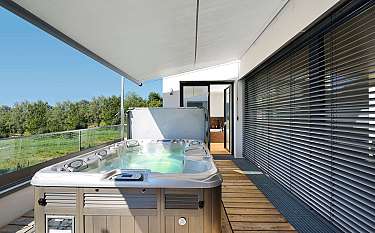 Ein Whirlpool auf einer überdachten Terrasse neben großen Fenstern und Blick ins Grüne.