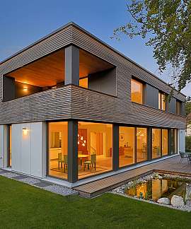 Modernes zweistöckiges Haus mit großer Glasfront, Terrasse und beleuchtetem Innenraum im Garten.