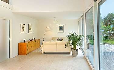 Helles Wohnzimmer mit Sofa, Pflanzen, Holzkommode und großen Glasfronten zum Garten.