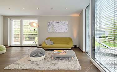 Heller Wohnraum mit gelbem Sofa, Stehlampe, Teppich und großen Fenstern mit Jalousien.