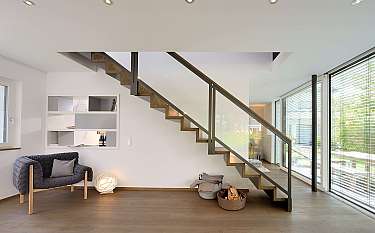 Heller Wohnraum mit offener Holztreppe, Sofa und großen Fensterfronten.