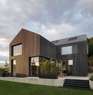 Modernes Haus mit vertikaler Holzfassade, großen Fenstern und Garten vor bewaldetem Hügel.
